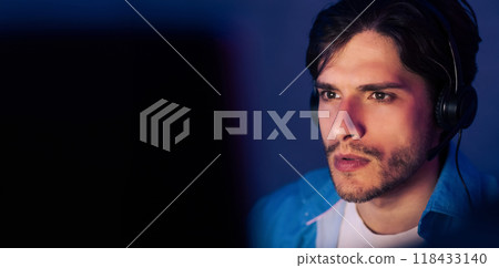 A young man with dark hair is wearing headphones and looking intently at a computer screen. He is wearing a blue jacket and a white t-shirt. The image is taken from a close-up perspective, copy space A young man with dark hair is wearing headphones and looking intently at a computer screen. He is wearing a blue jacket and a white t-shirt. The image is taken from a close-up perspective, copy space 118433140