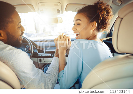 African American couple is sitting in the front seats of a car. The woman is looking over her shoulder with her mouth open in excitement and the man is looking forward, holding her hand 118433144
