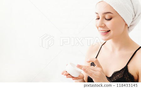 A cheerful young woman with a towel wrapped around her head is applying a creamy skincare product to her face. She is indoors, in a well-lit setting, and appears to enjoy her morning beauty routine. 118433145