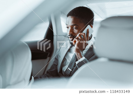 Black businessman in a suit sits in the back seat of a car and talks on his phone. The cars interior is clean and modern, and the man appears focused and professional. Black businessman in a suit sits in the back seat of a car and talks on his phone. The cars interior is clean and modern, and the man appears focused and professional. 118433149