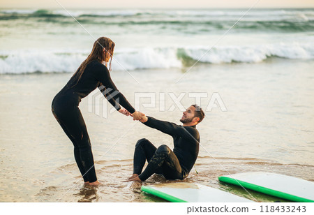 A woman in a wetsuit reaches out to help a man who has fallen off his surfboard in the shallows of a beach. He is sitting in the water with his surfboard nearby, while she is standing in the sand. 118433243
