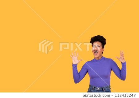 A young woman with short curly hair is standing in front of a yellow background. She is wearing a purple turtleneck and jeans and is screaming with her mouth wide open, copy space A young woman with short curly hair is standing in front of a yellow background. She is wearing a purple turtleneck and jeans and is screaming with her mouth wide open, copy space 118433247