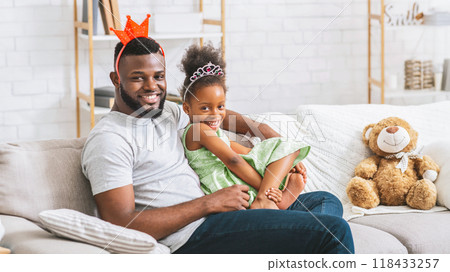 African American father and his young daughter sitting on a couch, smiling and looking at the camera. The girl is sitting on the fathers lap. They appear to be having a great time playing dress up 118433257