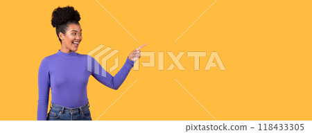 This is a photo of a young woman with dark skin and curly hair standing against a yellow background, wearing a purple long-sleeved shirt and blue jeans. She is smiling and pointing at copy space This is a photo of a young woman with dark skin and curly hair standing against a yellow background, wearing a purple long-sleeved shirt and blue jeans. She is smiling and pointing at copy space 118433305