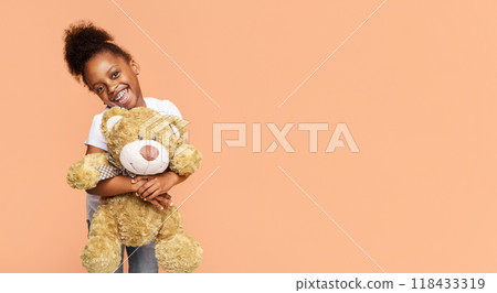 A little Black girl with curly hair smiles at the camera while holding a brown teddy bear against a peach-colored background, copy space A little Black girl with curly hair smiles at the camera while holding a brown teddy bear against a peach-colored background, copy space 118433319
