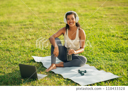 A young woman sits on a yoga mat in a park, wearing athletic clothing and headphones. She is smiling and holding a water bottle. Have online workout A young woman sits on a yoga mat in a park, wearing athletic clothing and headphones. She is smiling and holding a water bottle. Have online workout 118433328