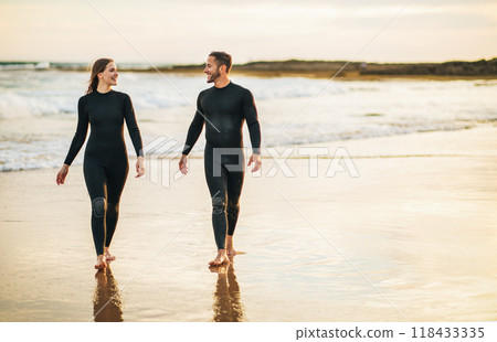 A couple in wetsuits walks along the sandy shore of a beach, with the golden light of sunset illuminating the scene. The man and woman are smiling and talking to each other, copy space 118433335