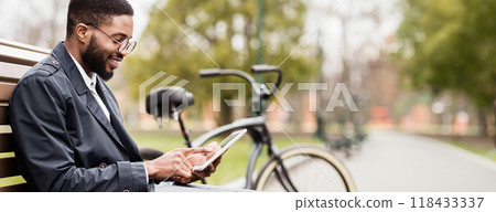 A professional black man is in a park, seated on a bench with a tablet and bicycle. The scene is relaxed and contemporary, capturing a moment of productivity in a natural setting. 118433337