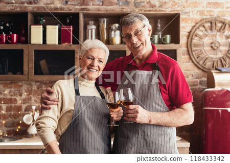 A senior couple, dressed in aprons, stand in a modern kitchen and toast with glasses of wine. They are smiling and looking at each other, clearly enjoying their time together. A senior couple, dressed in aprons, stand in a modern kitchen and toast with glasses of wine. They are smiling and looking at each other, clearly enjoying their time together. 118433342