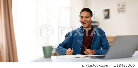 A young black student, wearing headphones, smiles while participating in an online class from his bright, cozy room. He sits at a desk with a laptop and notebook, focused on learning. 118433360