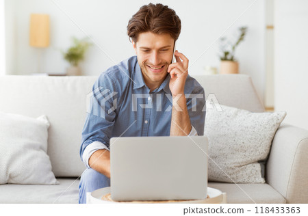 A man sits on a couch in his living room, using his laptop and talking on the phone. He is smiling and looking at the phone. 118433363