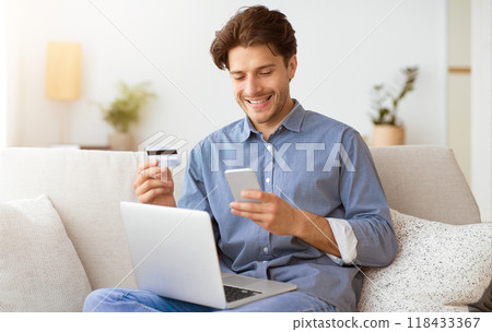 A man sits on a couch, smiling while looking at his smartphone. He holds a credit card in his other hand, and a laptop rests on his lap. 118433367