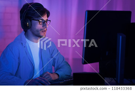 A young man with headphones and glasses is sitting at a computer in a room lit by purple and blue lights. He is looking at the monitor with a focused expression. 118433368