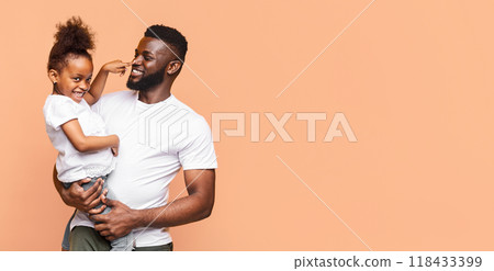 A happy black father is holding his daughter in his arms, while she playfully pokes his nose. They are both smiling, suggesting a strong bond between them, copy space 118433399