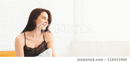 A young woman with long dark hair is sitting on a bed wearing a black camisole. She is smiling while looking out a window in a bright, sunlit bedroom. A young woman with long dark hair is sitting on a bed wearing a black camisole. She is smiling while looking out a window in a bright, sunlit bedroom. 118433400