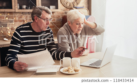 This image shows a senior couple sitting at a kitchen table, reviewing bills and a laptop together. They both have concerned expressions on faces, suggesting they are facing a financial challenge This image shows a senior couple sitting at a kitchen table, reviewing bills and a laptop together. They both have concerned expressions on faces, suggesting they are facing a financial challenge 118433414