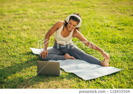 A woman with headphones is stretching her leg on a yoga mat outdoors in a park, while using a laptop. She is wearing a white sports bra and gray yoga pants. She is smiling while looking at the laptop. A woman with headphones is stretching her leg on a yoga mat outdoors in a park, while using a laptop. She is wearing a white sports bra and gray yoga pants. She is smiling while looking at the laptop. 118433504