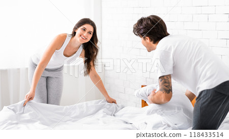 A woman in grey pants and a white tank top makes the bed while a man in white t-shirt and black pants watches. The couple are in a bedroom with white walls and a white brick wall. 118433514