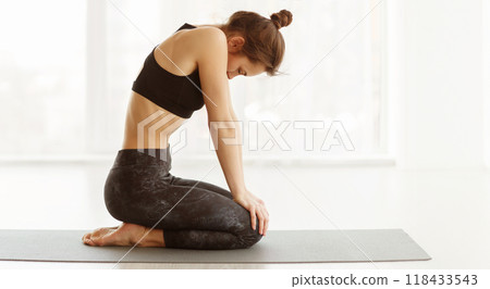 A woman is sitting on a yoga mat with her hands placed on her knees, engaging in a calm and focused posture. She appears to be in a peaceful state of concentration and relaxation. 118433543