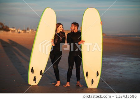 A young couple stands on a sandy beach at sunset, holding their surfboards. The sun is setting in the background, and the sky is a beautiful orange and pink. 118433596