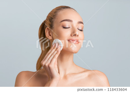 A young woman with long brown hair, wearing no makeup, is applying a cotton pad to her cheek. She has closed eyes and is gently rubbing the cotton pad in a circular motion 118433604