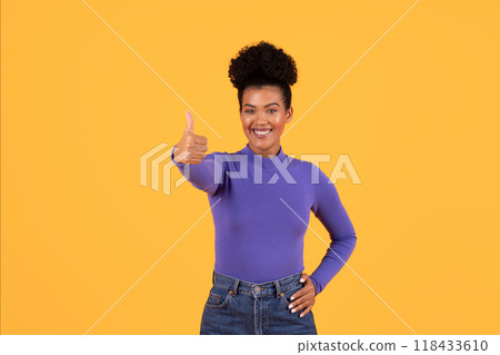 A smiling woman in a purple shirt stands against a yellow backdrop and gives a thumbs up with her right hand. She has dark brown skin, dark curly hair, and is wearing blue jeans. 118433610