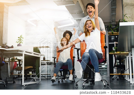 Funny working time. Overjoyed millennial colleagues racing on office chairs in empty coworking space 118433628