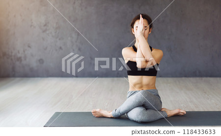 A young woman sits cross-legged on a yoga mat in a tranquil room, her hands in the Anjali Mudra pose. She appears calm and focused, exhibiting a balance of mind and body, copy space A young woman sits cross-legged on a yoga mat in a tranquil room, her hands in the Anjali Mudra pose. She appears calm and focused, exhibiting a balance of mind and body, copy space 118433633