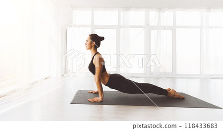 A woman is engaged in a yoga session, performing the upward-facing dog pose on a black mat in a well-lit, airy studio. The environment suggests serenity and the pursuit of wellness 118433683
