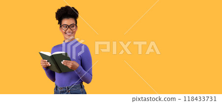 A young woman with short, curly hair is wearing glasses and a purple turtleneck sweater and is smiling while reading a book. She is standing against a solid yellow background. 118433731