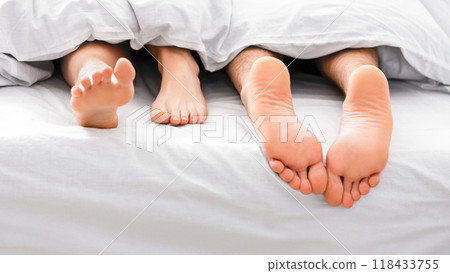 A close-up image of two bare feet, one male and one female, sticking out from underneath white bedding, suggesting a couple is sleeping in bed. A close-up image of two bare feet, one male and one female, sticking out from underneath white bedding, suggesting a couple is sleeping in bed. 118433755