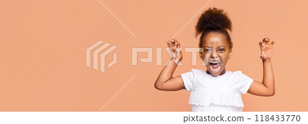 A little girl with curly hairstyle is standing in front of a peach-colored background, shouting with her arms raised in the air. Her expression is intense, and she is looking directly at the camera. A little girl with curly hairstyle is standing in front of a peach-colored background, shouting with her arms raised in the air. Her expression is intense, and she is looking directly at the camera. 118433770