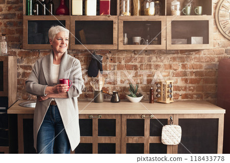 A senior woman stands in a rustic kitchen with exposed brick walls, holding a red mug of coffee in her hands. She is smiling as she looks out of the window, enjoying her morning drink, copy space 118433778