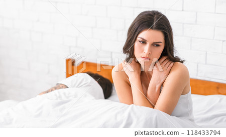 A woman sits in bed with a forlorn expression, looking down at her hands. Her partner sleeps soundly beside her. The image conveys a sense of sadness, loneliness, and emotional distance. 118433794