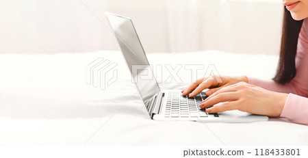 A woman is sitting on a bed, focused on using a laptop computer. She is typing and browsing online content, cropped, panorama 118433801