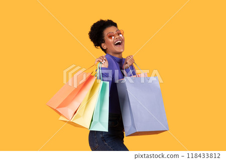 A woman with curly hair and heart-shaped sunglasses is smiling widely and holding several colorful shopping bags. She is standing against yellow background, suggesting a joyful shopping experience. 118433812