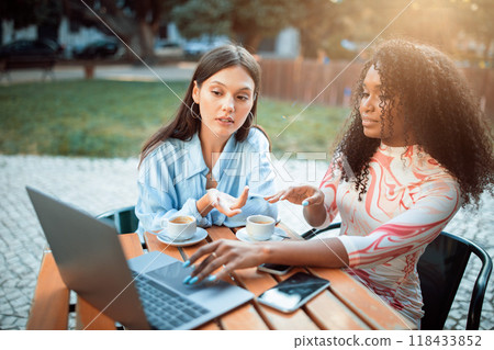 Two women are seated at a table outside a cafe. One woman is using a laptop computer while the other woman gestures and looks on. There is a cell phone on the table next to the laptop. 118433852