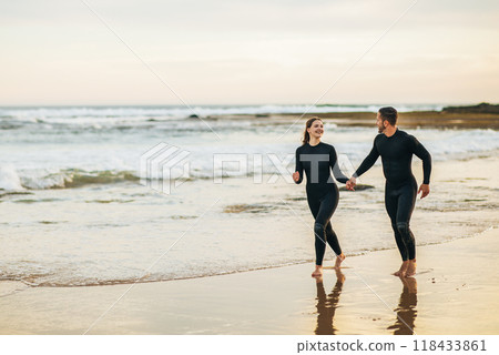 A happy couple runs along the sandy beach holding hands, both wearing black wetsuits. The ocean waves gently lap at the shore behind them as the sun sets in the distance, copy space A happy couple runs along the sandy beach holding hands, both wearing black wetsuits. The ocean waves gently lap at the shore behind them as the sun sets in the distance, copy space 118433861