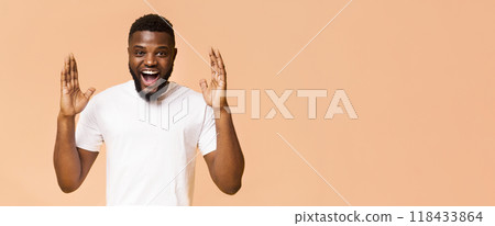 A young black man wearing a white t-shirt smiles and looks shocked while raising his hands in the air. He has a light brown complexion and a full beard. He is standing in front of a peach background. 118433864