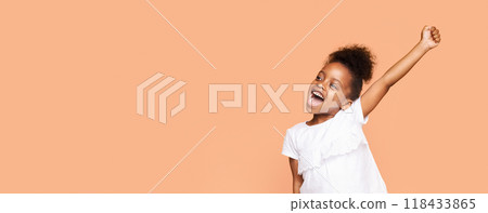 A little black girl with curly hair and a bright smile raises her arm in celebration against a peach-colored background. She is wearing a white t-shirt and appears to be full of joy and excitement A little black girl with curly hair and a bright smile raises her arm in celebration against a peach-colored background. She is wearing a white t-shirt and appears to be full of joy and excitement 118433865