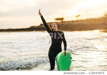 A woman wearing a black wetsuit smiles and holds a green surfboard in the ocean. The water is shallow and the sun is setting in the background, casting a warm glow on the scene. 118433875