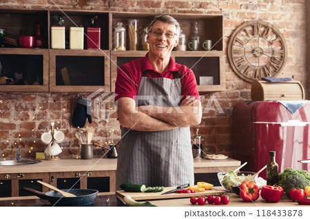An older man stands in his kitchen, arms crossed, wearing a gray apron over a red shirt. He is smiling warmly and looks ready to cook. The kitchen features a brick wall, a wooden countertop An older man stands in his kitchen, arms crossed, wearing a gray apron over a red shirt. He is smiling warmly and looks ready to cook. The kitchen features a brick wall, a wooden countertop 118433878