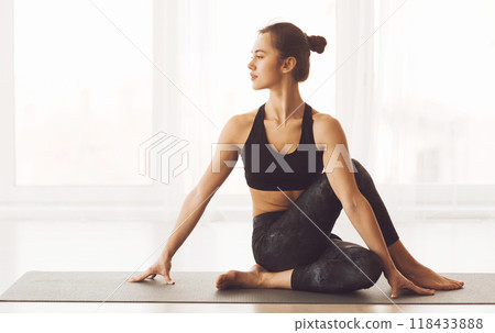 A young woman is captured in a serene moment while engaged in a yoga pose, calmly stretching her body on a mat within a brightly lit room, suggesting an atmosphere of contemplation and wellness. 118433888