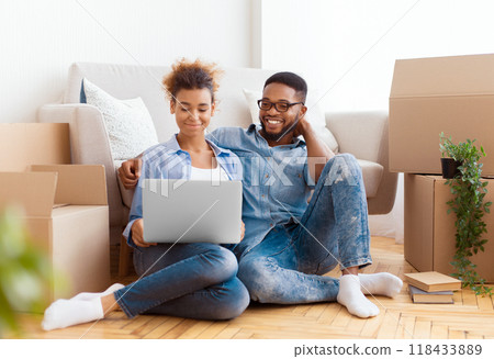 Relocation Concept. Spouses Browsing On Laptop Ordering Furniture For New Apartment Sitting On Floor Among Moving Boxes Indoor. Selective Focus Relocation Concept. Spouses Browsing On Laptop Ordering Furniture For New Apartment Sitting On Floor Among Moving Boxes Indoor. Selective Focus 118433889
