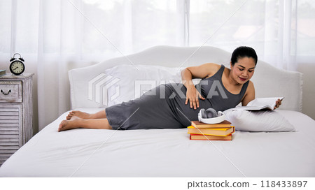 One Pregnant woman lying on bed reading book with stack of books and headphones One Pregnant woman lying on bed reading book with stack of books and headphones 118433897