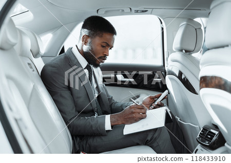 African American businessman sits in the backseat of a car, working on a notepad and using a smartphone. He is dressed in a suit and tie, and appears to be focused on his task. 118433910
