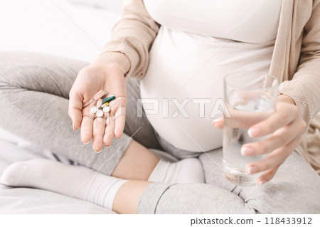 A pregnant woman is seen holding a pill in one hand and a glass of water in the other, preparing to take medication. The image shows the importance of taking prescribed medication during pregnancy 118433912