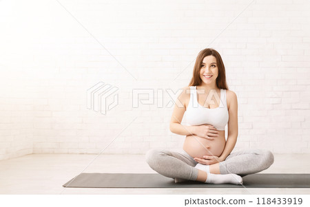 A pregnant woman is seated on a yoga mat, engaged in a prenatal yoga session. She is focusing on gentle stretches and breathing exercises to support her changing body and prepare for childbirth. 118433919