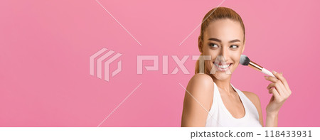 This photo shows a young woman with a bright smile applying blush to her cheek with a makeup brush. She is wearing a white tank top and has her hair pulled back in a ponytail, copy space This photo shows a young woman with a bright smile applying blush to her cheek with a makeup brush. She is wearing a white tank top and has her hair pulled back in a ponytail, copy space 118433931