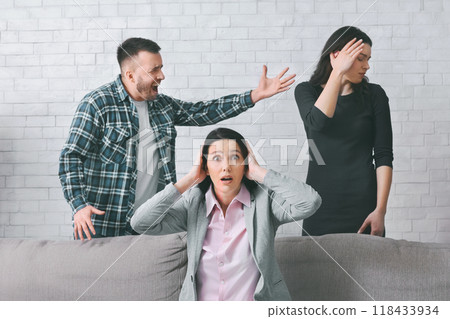A woman sits on a couch with her hands over her ears as a man yells at another woman standing behind the couch. Woman standing behind the couch with her hand on her forehead. A woman sits on a couch with her hands over her ears as a man yells at another woman standing behind the couch. Woman standing behind the couch with her hand on her forehead. 118433934